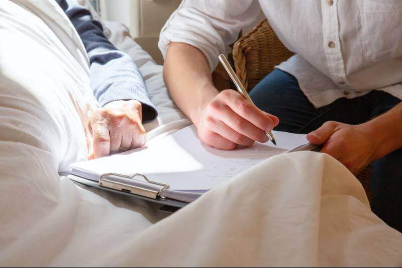 man writing on a form beside a elderly person's bed